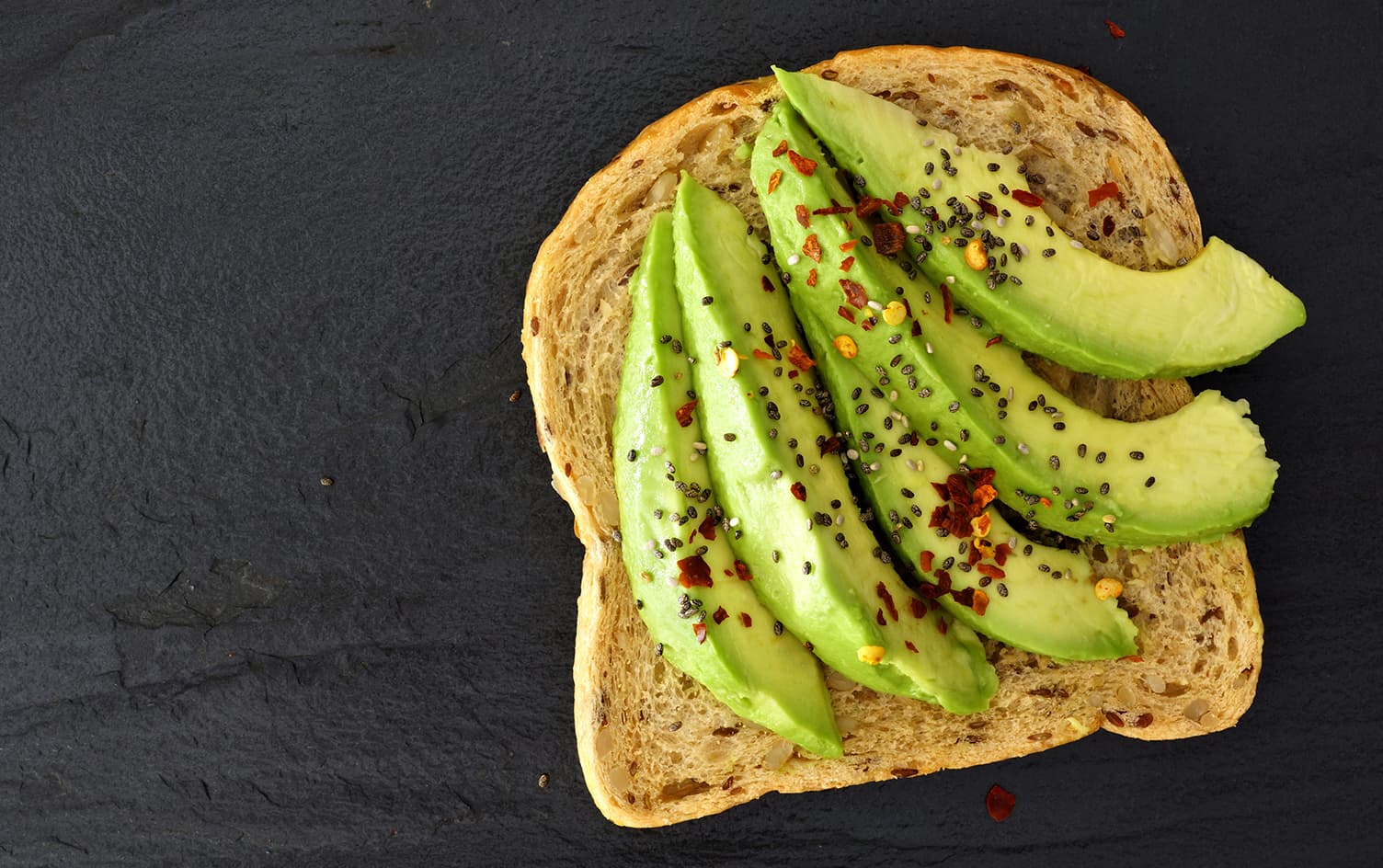 A slice of whole grain toast topped with sliced avocado, sprinkled with black chia seeds and chili flakes for added fiber, placed on a dark slate surface. MyFitnessPal Blog